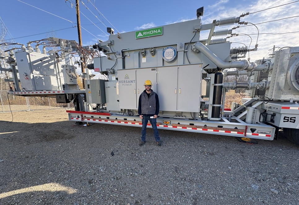 A man in a hard hat stands next to a large, gray electrical transformer on a trailer.